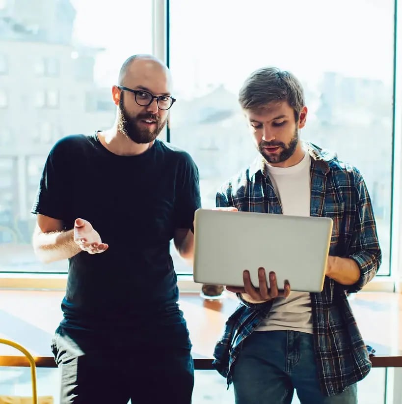 Two men stand by a window, engaged in discussion. One, in glasses and a black shirt, gestures while the other, in a plaid shirt, holds a laptop. Natural light fills the room