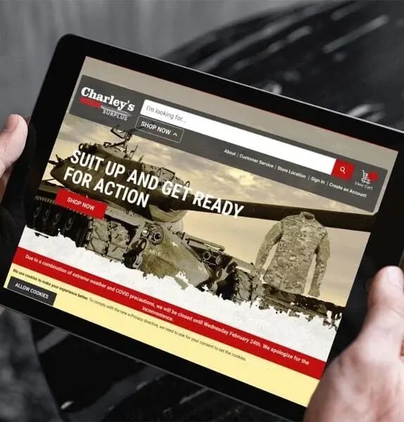 A person holds a tablet displaying an online store for military gear. The screen shows a tank, a camouflage jacket, and the text _Suit up and get ready for action_