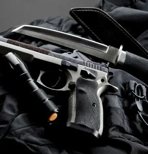 A tactical setup displaying a black handgun, a combat knife with a sheath, and a flashlight laid on textured black fabric. The scene conveys readiness