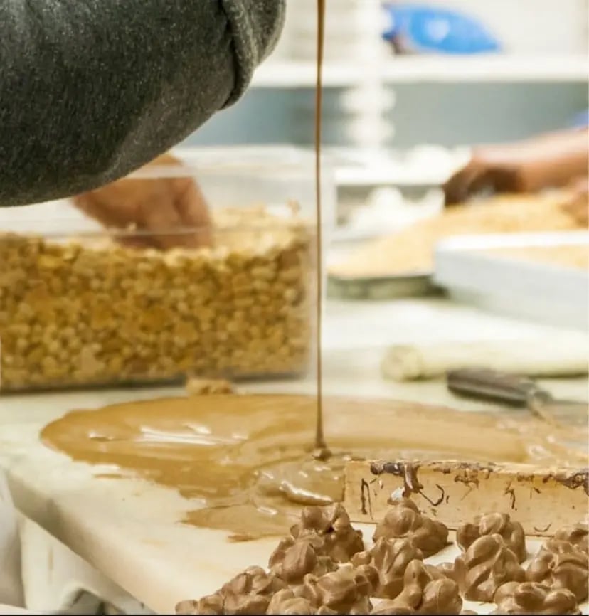 Chocolate being poured over clusters of nuts on a confectionery production line, with trays of ingredients in the background.