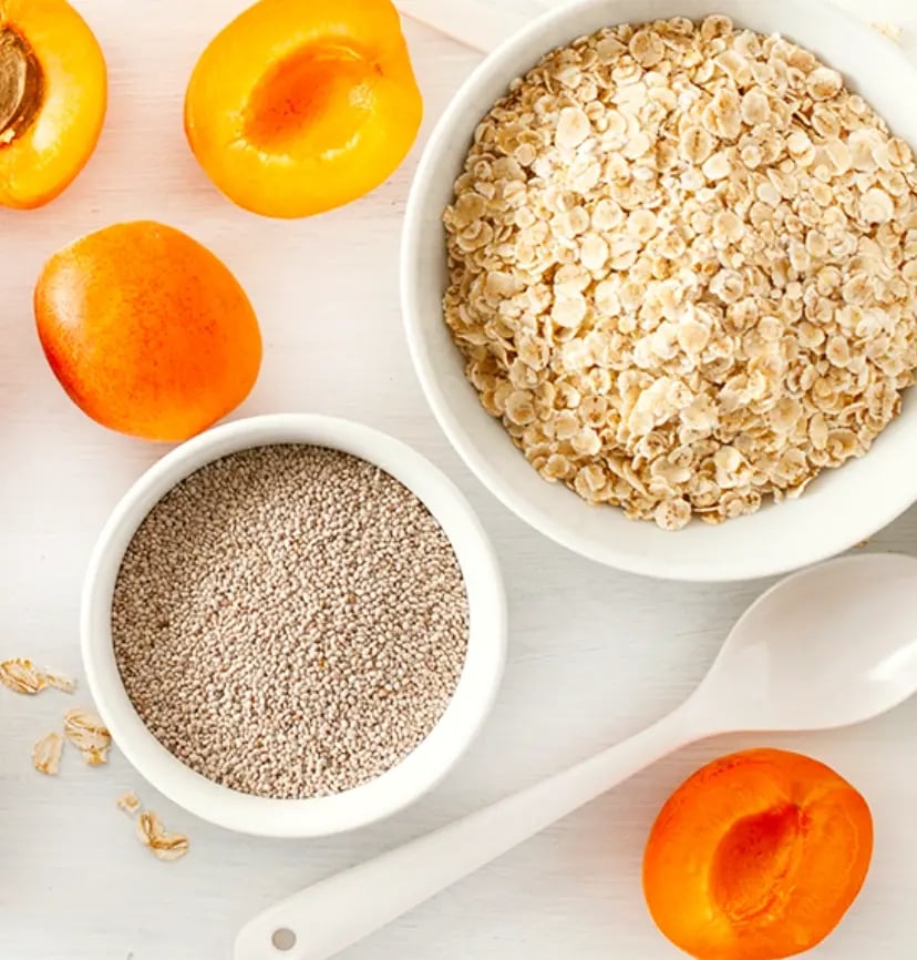 Top view of rolled oats, quinoa seeds, and fresh apricots in bowls with a spoon on a light background, showcasing healthy breakfast ingredients.