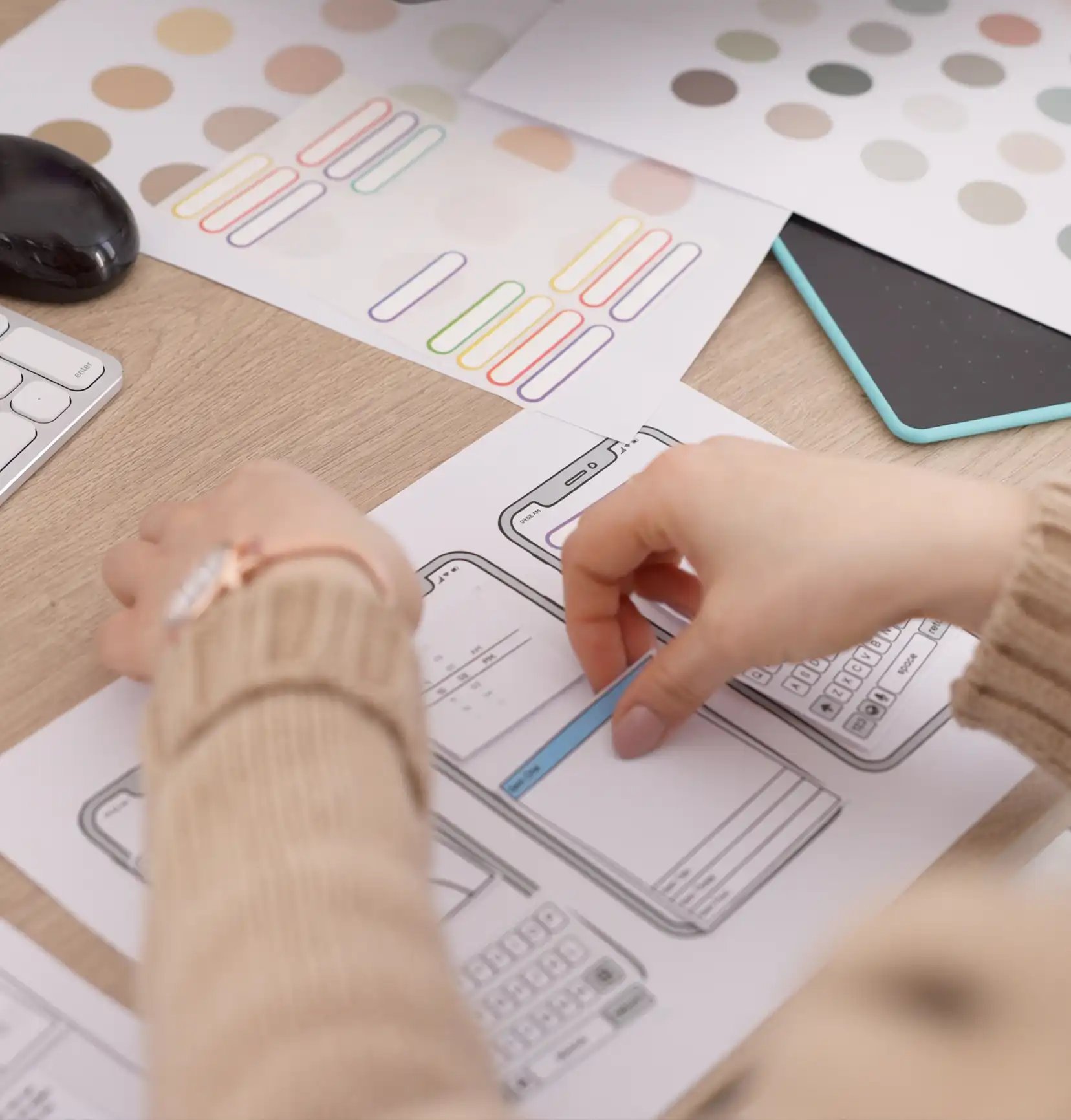 Hands are sketching mobile app designs on paper, surrounded by color palettes and UI templates on a desk, conveying creativity and focus.