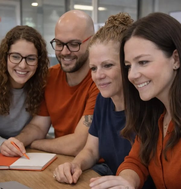 Four professionals collaborate at a modern office desk, leaning forward and smiling as they review work on a computer, with notebooks and burnt orange (#CB6A52) accents creating a warm, cohesive workspace aesthetic.