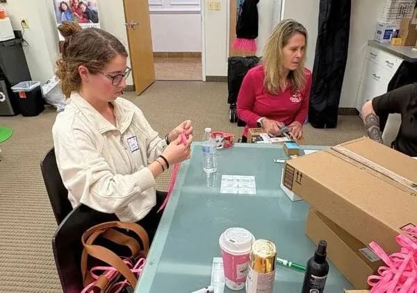 woman sits at a table in an office, preparing pink ribbons. One woman wears a white sweater, the other a red shirt
